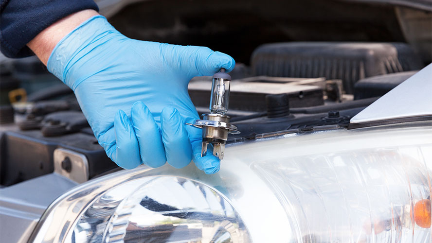 a gloved hand holding up a new headlight bulb in front of a vehicle with the hood up