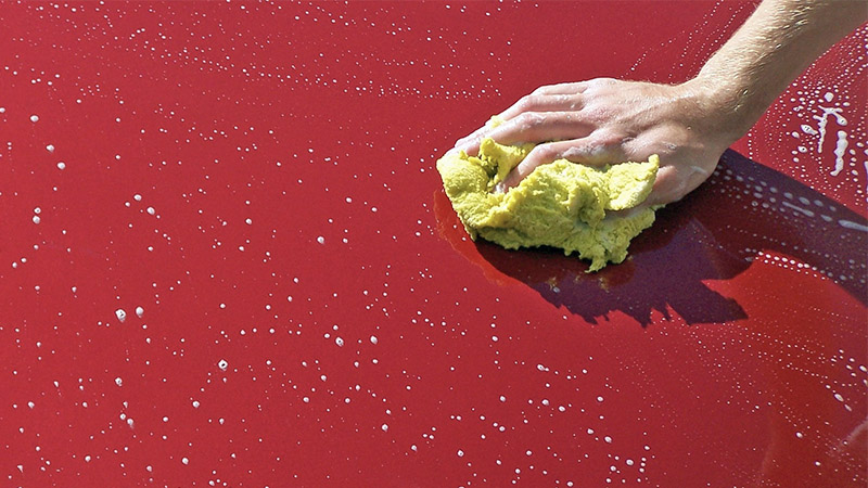 a hand holding a yellow sponge washes a red car