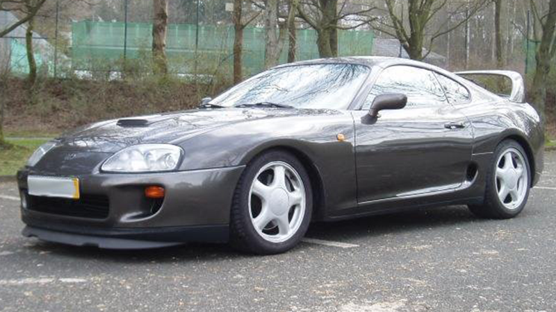A grey Toyota Supra in a parking lot with trees behind it.