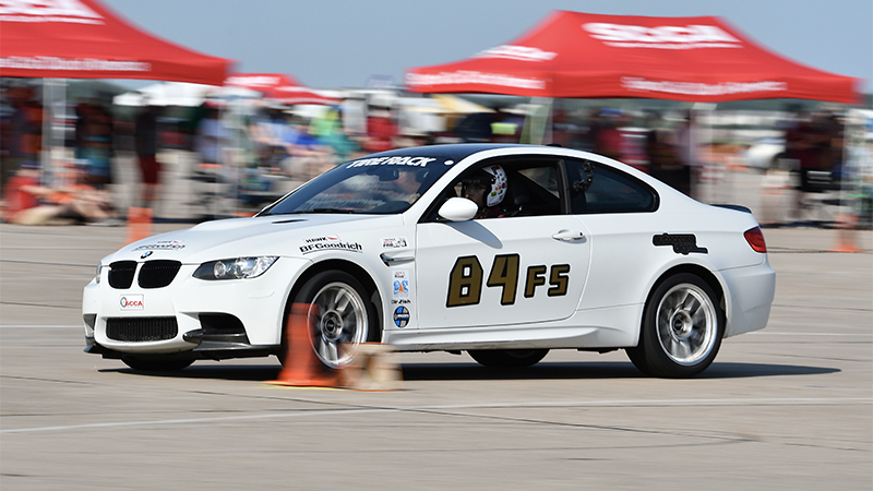 A white BMW on an autocross course with cones marking the track and red tents in the background 