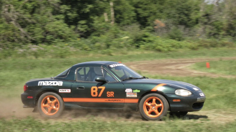 a green Miata on a dirt track carved out of a grassy field with trees in the background