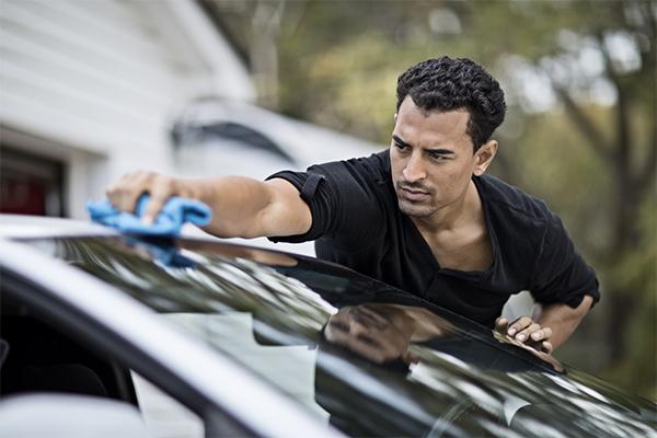 a man in a short-sleeved shirt reaches across the top of the windshield with a blue cloth for cleaning